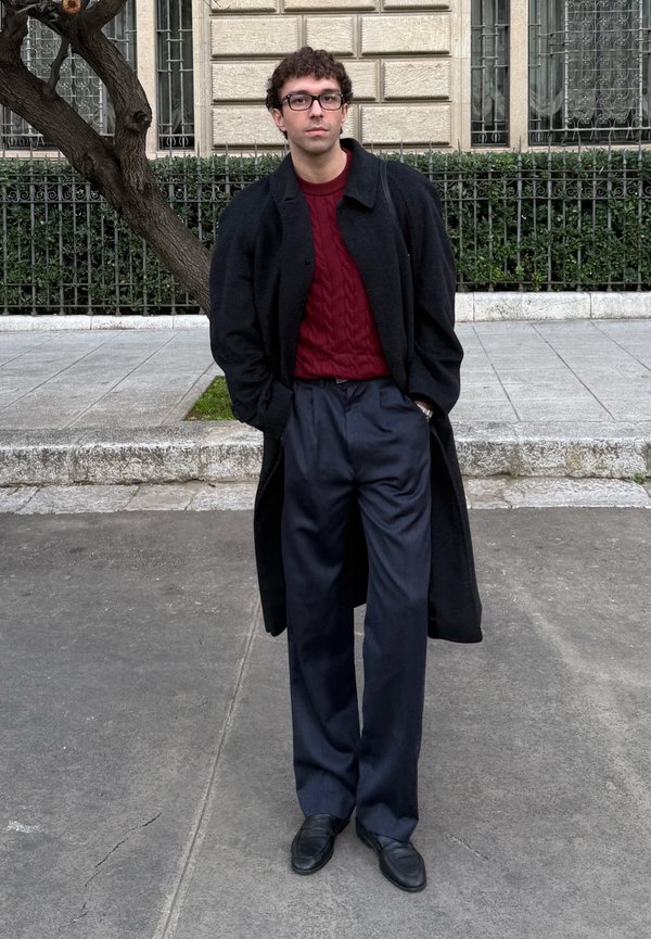 Giovane uomo con capelli ricci e occhiali che indossa un cappotto nero, un maglione rosso, pantaloni blu navy e scarpe nere, in piedi su un marciapiede cittadino.