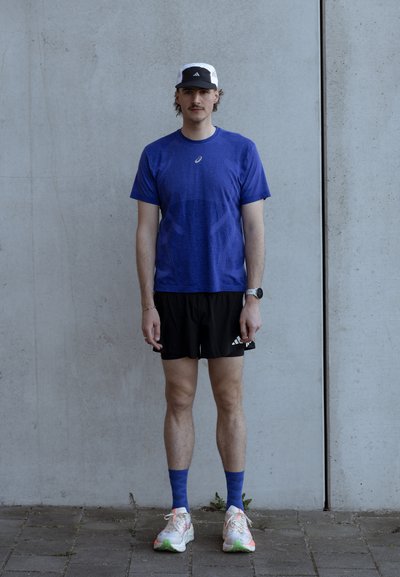 Joven con camiseta deportiva azul, pantalones cortos negros, calcetines azules, zapatillas de correr blancas y gorra en blanco y negro, apoyado contra una pared de hormigón.