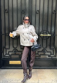 Beżowy wełniany płaszcz typu peacoat z podwójnym zapięciem na guziki i dużymi guzikami, w połączeniu z dopasowanymi brązowymi spodniami i brązowymi butami na obcasie z ostrym noskiem. Czarna torebka.