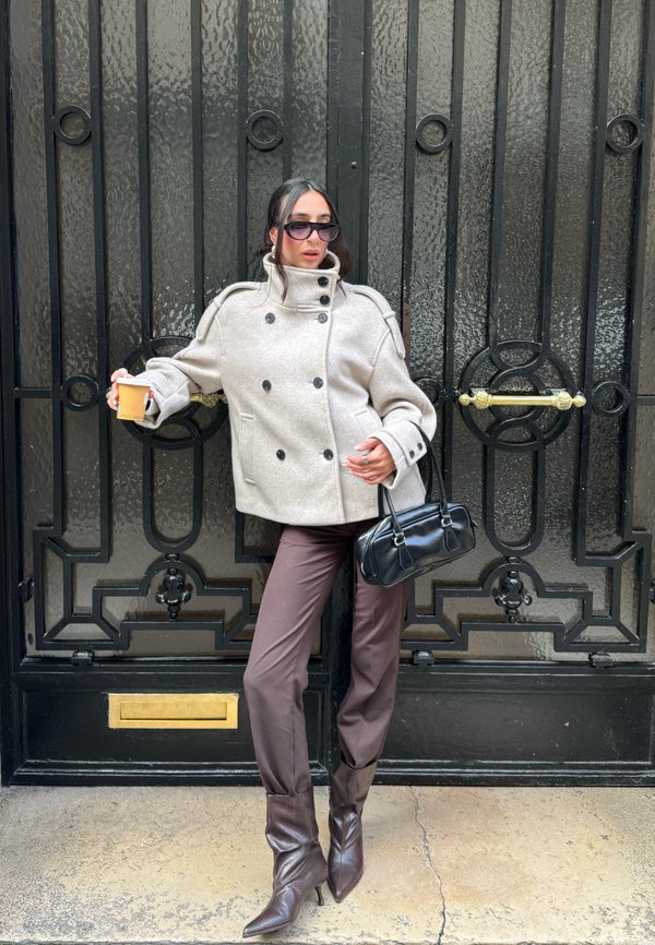 Beige uldpeacoat med dobbeltradet design og store knapper, kombineret med tilpassede brune bukser og spidse brune støvler med hæl. Sort håndtaske.