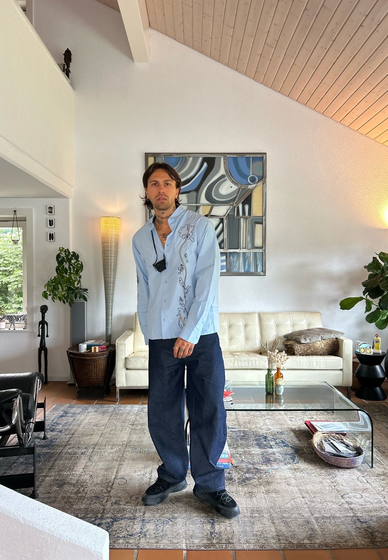 Uomo con capelli neri che indossa una camicia azzurro chiaro e pantaloni scuri, in piedi in un soggiorno moderno con divano bianco, tavolo di vetro e arte murale astratta.