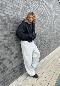 Czarna kurtka bomberka z kapturem wykończonym sztucznym futrem, noszona na jasnoszarych spodniach dresowych. Ciemne buty, stojące na tle teksturowanej szarej ceglanej ściany.