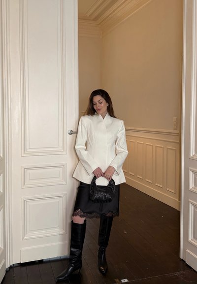 Mujer con blazer blanco, falda negra con borde de encaje y botas negras hasta la rodilla, de pie sosteniendo un bolso negro tejido junto a unas puertas dobles blancas cerradas.