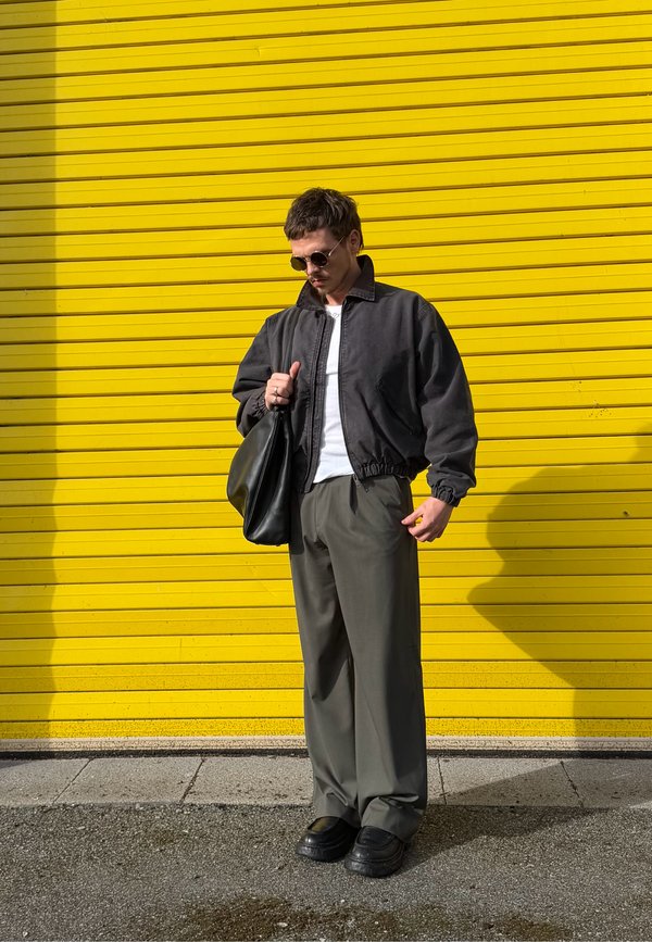 Hombre con gafas de sol, chaqueta negra, camisa blanca y pantalones verdes que sostiene una bolsa negra, de pie frente a una pared de metal corrugado amarilla.