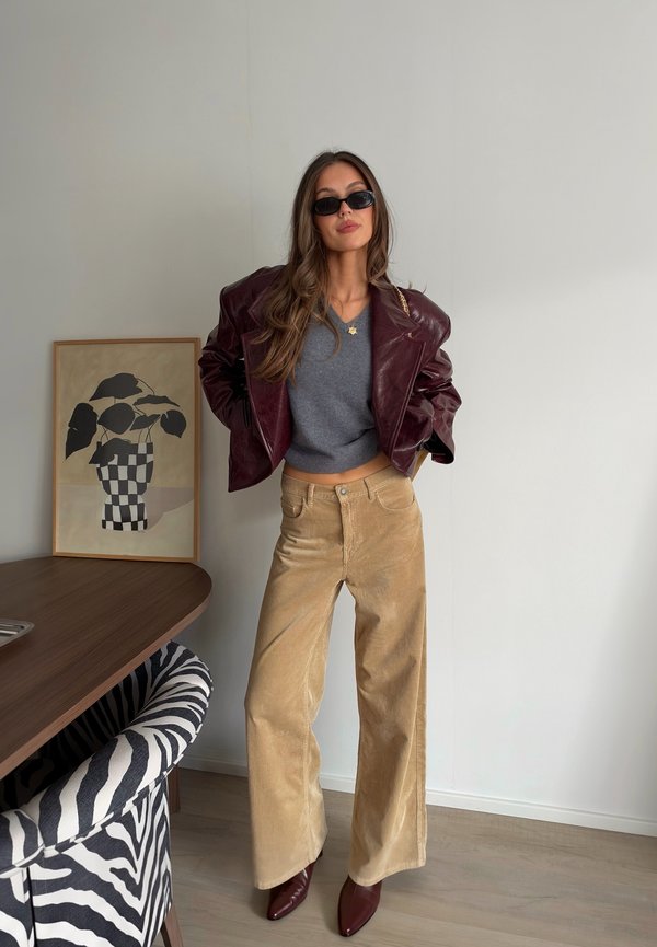 Une femme pose à l’intérieur, portant des lunettes de soleil, une veste en cuir bordeaux, un haut gris, un pantalon ample beige et des bottes pointues assorties.