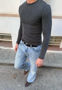 Άνδρας που φοράει εφαρμοστή σκουρόγρια μακρυμάνικη μπλούζα, ανοιχτόχρωμο μπλε τζιν με μαύρη ζώνη και καφετιά μυτερά παπούτσια, στέκεται σε πεζοδρόμιο.