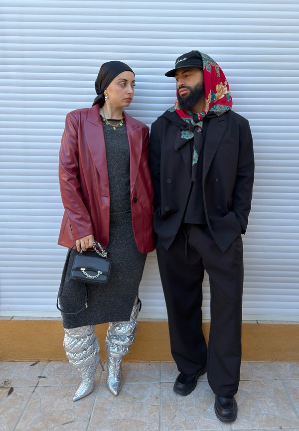 Dos personas están juntas: una lleva una blazer de cuero roja sobre un vestido gris y botas plateadas acolchadas hasta la rodilla; la otra viste un traje negro con una bufanda colorida.