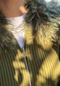 Cardigan verde măsliniu cu dungi și guler pufos, cu fermoar argintiu. Tricou alb vizibil dedesubt, adăugând un contrast de texturi.