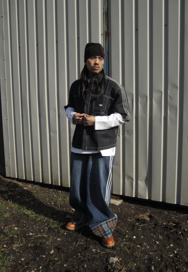 Homme aux cheveux longs portant un bonnet noir, une veste noire superposée, un pantalon bleu large et des bottes marron, debout contre un mur en métal ondulé.
