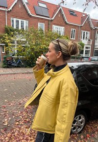 Kvinne i gul jakke som drikker kaffe, står på en løvdekket gate med mursteins hus, en svart bil og sykler i bakgrunnen.