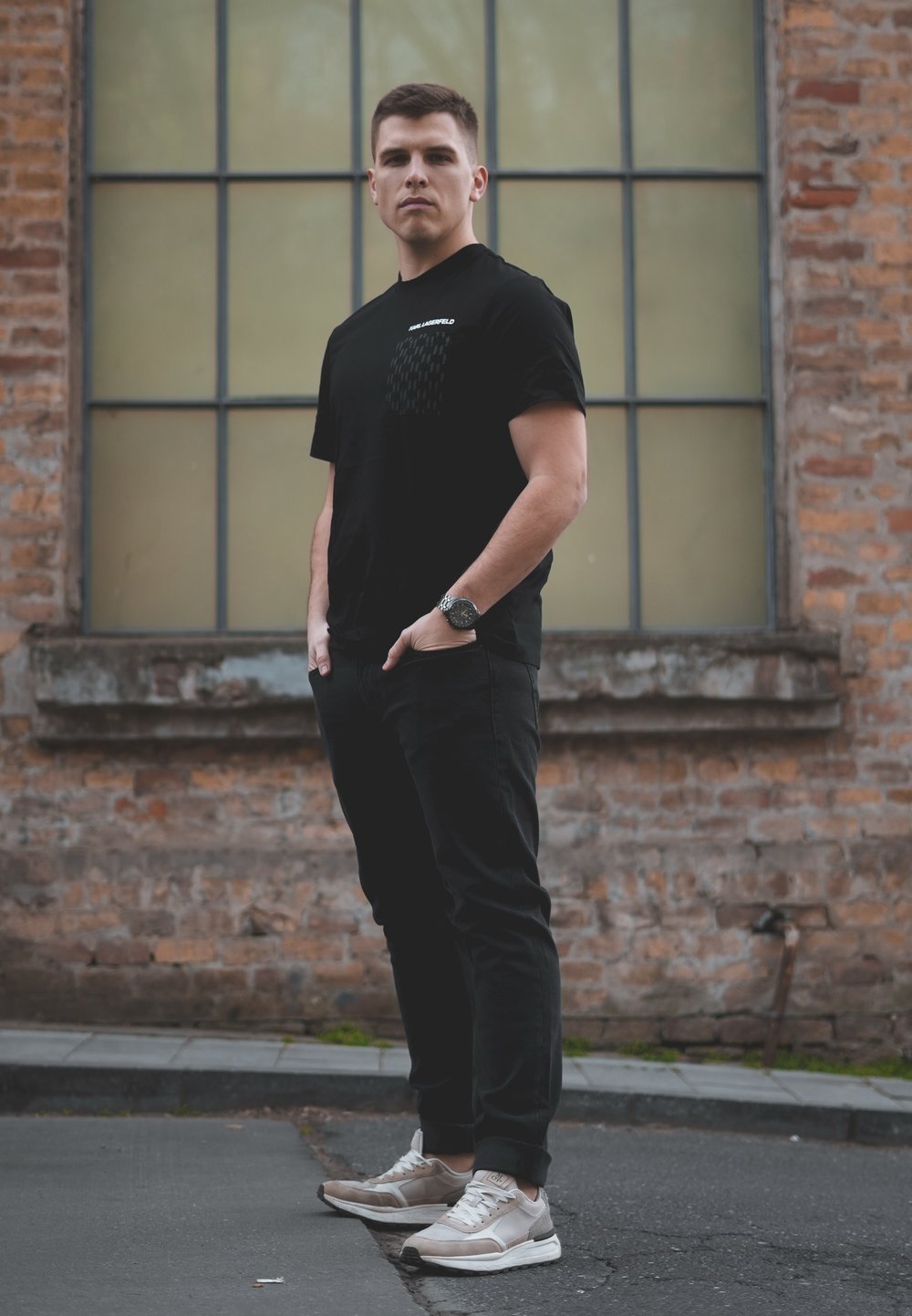 Melna t-krekls ar teksturētu kabatu, melnas bikses un gaišas kedas. Stāv pret ķieģeļu sienu ar lieliem zaļgani nokrāsotiem logiem.