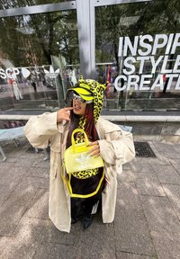 Rumena kapuca s leopardskim vzorcem in ušesi, bež oversize plašč, črne hlače in rumena torbica Adidas. Jasna sončna očala z drznim dizajnom.