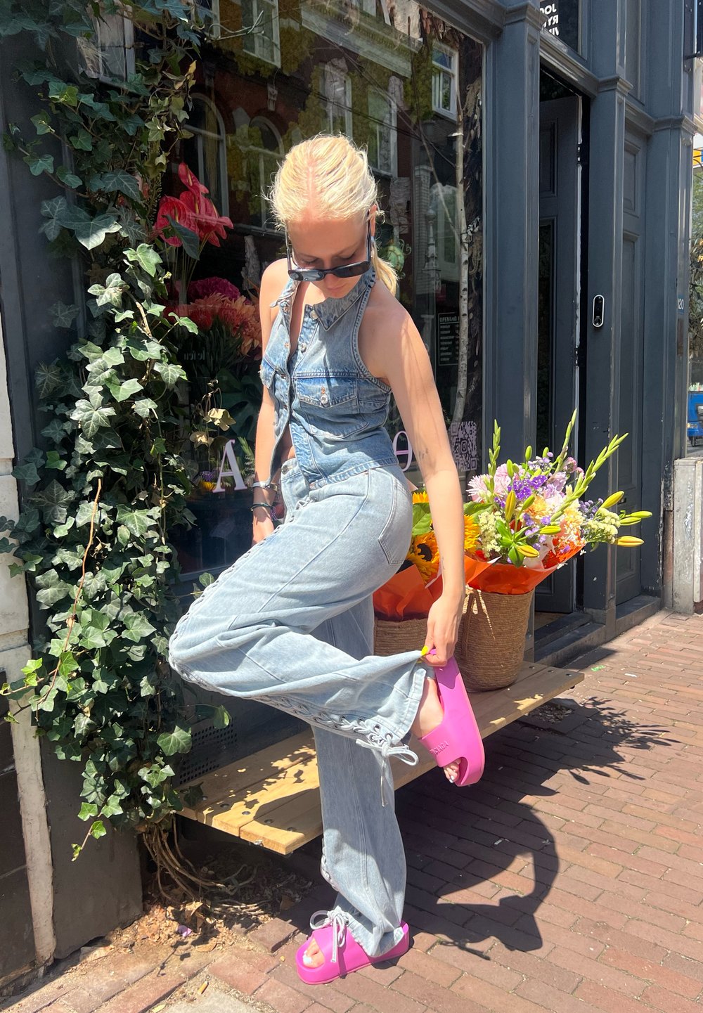 Denim overalls in light blue, featuring a button front and loose fit, paired with bright pink platform sandals. Green ivy and flowers in the background.