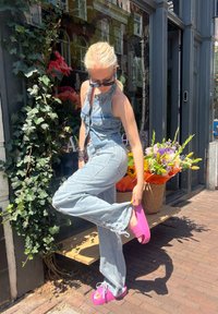 Denim overalls in light blue, featuring a button front and loose fit, paired with bright pink platform sandals. Green ivy and flowers in the background.