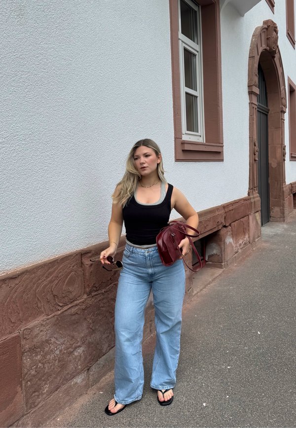 Mladá žena kráča vedľa bielej budovy s červeným lemovaním, oblečená v čiernom tielku, svetlomodrých džínsoch, žabkách, drží slnečné okuliare a červenú tašku.