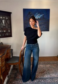 Femme en haut noir et jean à jambes larges tenant une chaise en bois antique dans une pièce avec un bureau en bois et une peinture bleue.