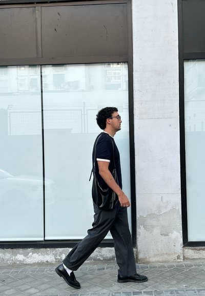 Hombre con cabello rizado, camiseta negra, pantalones grises y zapatos negros caminando past un edificio con grandes ventanales de vidrio y una pared blanca.