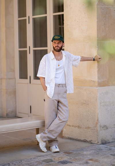 Camisa blanca de manga corta con un patrón de rombos, camiseta blanca sin mangas, pantalones beiges y zapatillas blancas. Accesorios como una gorra verde y un collar.