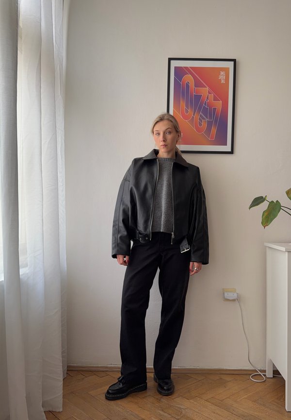 Jeune femme portant une veste en cuir noire, un pull gris, un pantalon noir et des chaussures noires, se tenant à l'intérieur près d'une fenêtre et d'une plante.