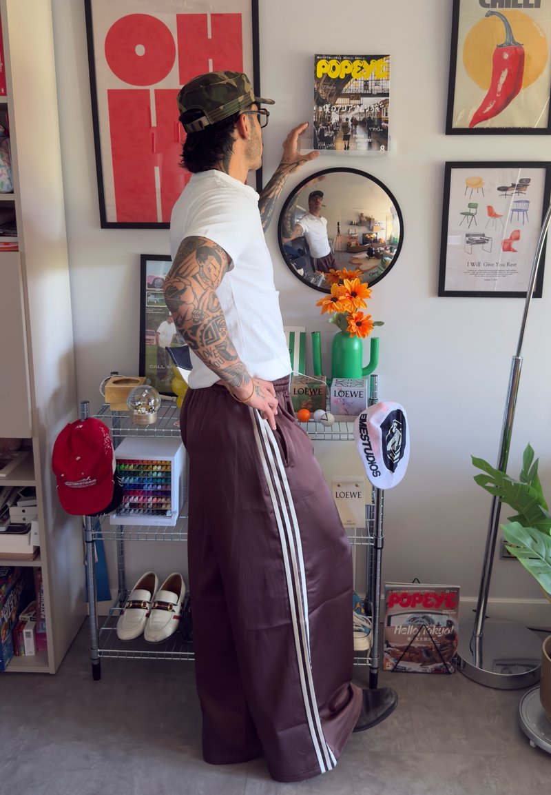 Braune weit geschnittene Hosen mit weißen Streifen, ein weißes T-Shirt und eine Camouflage-Mütze. Im Hintergrund ein Spiegel, bunte Aufbewahrung und dekorative Blumen.