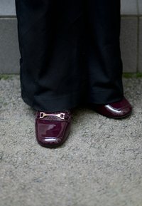 Pantofi din piele lăcuită burgundy cu vârfuri pătrate, accentuați de o cataramă aurie. Purtați cu pantaloni din țesătură neagră. Solul are o textură.
