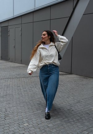 Weißer Reißverschluss-Sweatshirt, hellblaue weite Jeans, schwarze Loafers, läuft auf Kopfsteinpflaster. Lange dunkle Haare, minimalistischer schwarzer Handtasche.