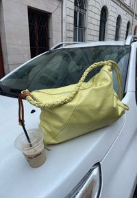Sac à main en cuir jaune avec une poignée tressée et une fermeture éclair, posé à côté d'un café glacé sur une surface blanche.