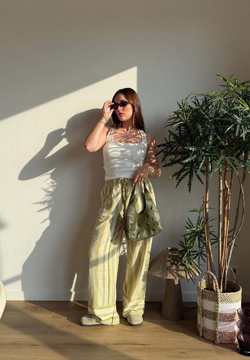 Femme portant des lunettes de soleil, un débardeur blanc, un pantalon large rayé, tenant un sac à main vert, debout près d'une grande plante en pot dans la lumière du soleil à l'intérieur.