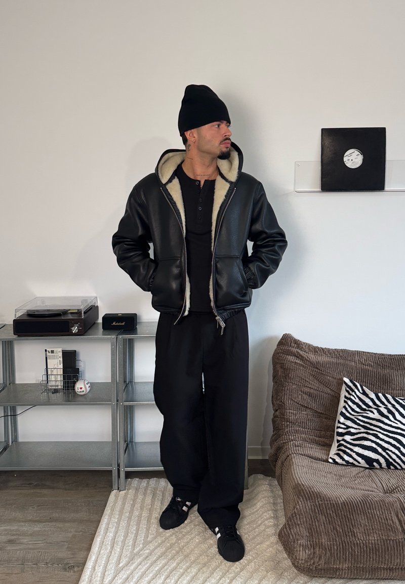 Sort læderjakke med cremefarvet shearlingfor, sort henleyskorte, løse sorte bukser og sorte sneakers mod en neutral baggrund.