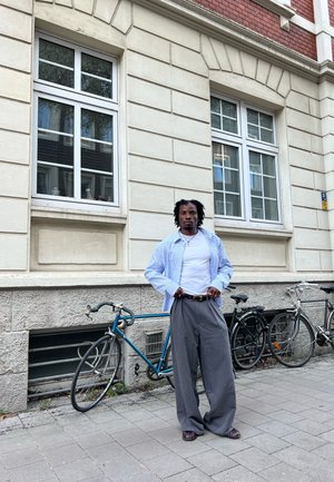 Camisa de manga larga azul claro sobre una camiseta blanca, pantalones grises de pierna ancha y sandalias marrones. Apoyado contra una bicicleta azul.
