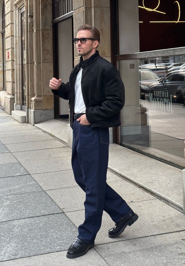 Hombre con chaqueta negra, camisa blanca, pantalones oscuros y gafas caminando por la acera de la ciudad cerca de la entrada de un bar-cafetería.