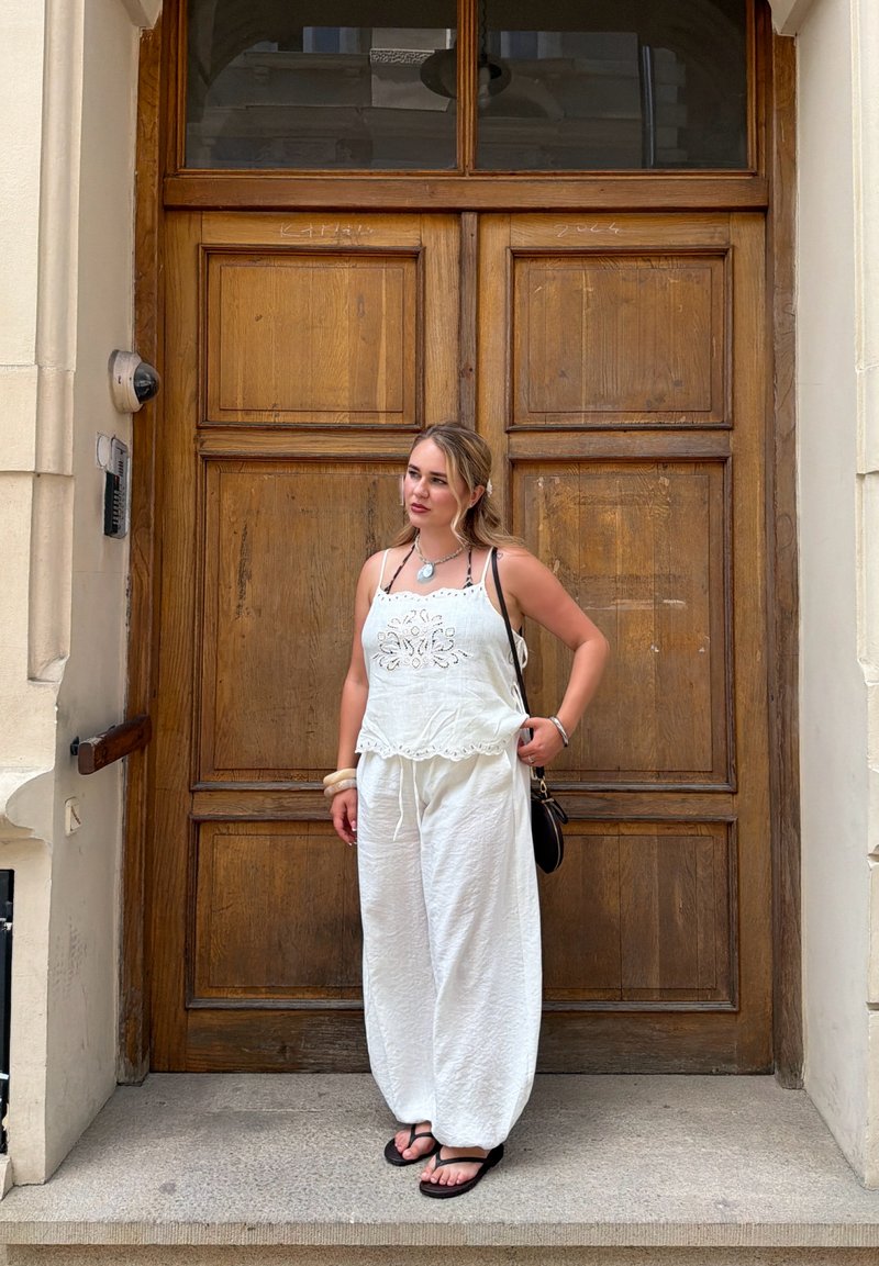 Jeune femme en haut blanc sans manches et pantalon se tient sur des marches en pierre devant une grande porte en bois. Elle porte des sandales noires et porte un sac noir.