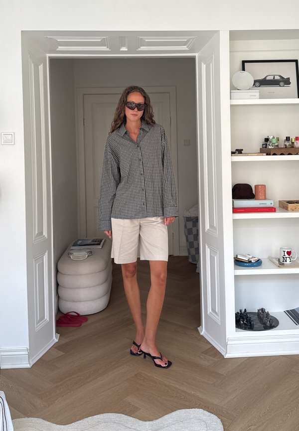 Femme portant une chemise grise à carreaux, un short beige, des sandales noires et des lunettes de soleil, debout dans une porte d'une pièce minimaliste.
