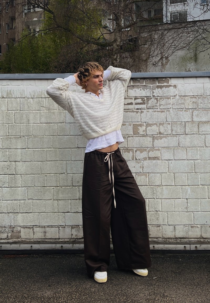 Jeune personne aux cheveux bouclés posant les mains derrière la tête, portant un pantalon ample foncé, des baskets blanches, une chemise blanche et un pull à rayures beige en plein air.