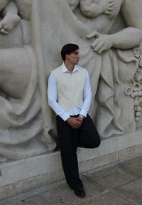 Bela brezrokavna jakna preko dolgih rokavov bele srajce, v paru z temnimi hlačami s črtami. Stoječa ob teksturiranem kamenem zidu.