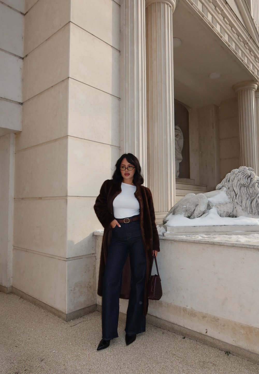 Femme en manteau sombre en fourrure, haut blanc, jean taille haute foncé et lunettes, debout près d'un bâtiment en pierre classique avec une colonne et une statue de lion.