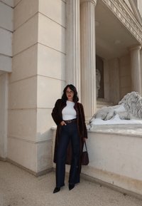 Femme en manteau sombre en fourrure, haut blanc, jean taille haute foncé et lunettes, debout près d'un bâtiment en pierre classique avec une colonne et une statue de lion.