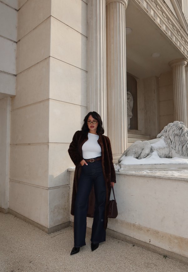 Kvinde iført mørk pelsfrakke, hvid top, mørke jeans med høj talje og briller står ved siden af en klassisk stenmuret bygning med søjle og løvestatue.