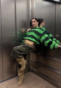 Groen en zwart gestreepte oversized shirt, camouflage broeken en beige stiletto laarzen. Accentdetails omvatten een ronde tan riemtas en een metalen liftachtergrond.
