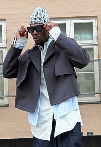 Lyhyt tumman takin päällä oleva siniraitainen paita, jota täydentää kuvioitu neulottu hattu ja mustat aurinkolasit, tekstuurilla varustettu kaupunkitausta.