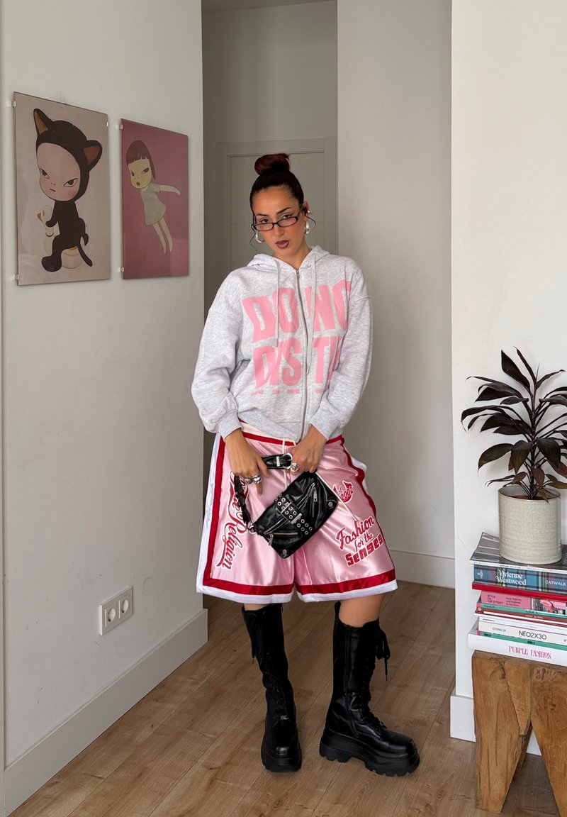 Femme portant des lunettes, un sweat à capuche gris, un short rose et rouge, tenant un sac noir, debout à l'intérieur près d'une œuvre d'art et d'une plante posée sur des livres.