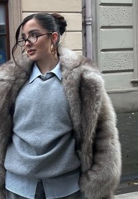 Sivá mikina cez modrú pruhovanú košeľu, doplnená béžovým kabátom z umelého kožušiny. Ozdobená okrúhlymi zlatými kruhovými náušnicami a okuliarmi.