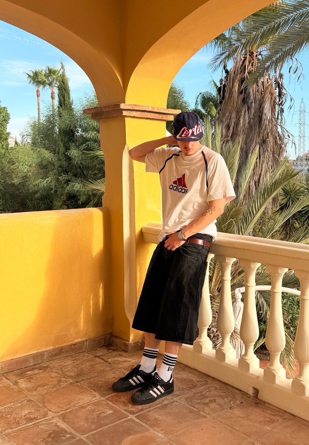 Svarta denimshorts, vit Adidas-t-shirt med röd logotyp, svart keps med vit text, och svarta Adidas-skor med vitrandiga strumpor.