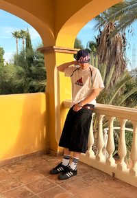 Svarta denimshorts, vit Adidas-t-shirt med röd logotyp, svart keps med vit text, och svarta Adidas-skor med vitrandiga strumpor.