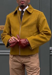 Άνδρας που φοράει μπουφάν με φερμουάρ σε μουσταρδί κίτρινο, μπεζ παντελόνι, σκούρα γραβάτα, λευκό πουκάμισο και φοράει σκουτεκό κόκκινα δερμάτινα γάντια.