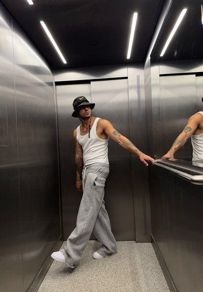 Una persona está de pie en un ascensor vestida con una camiseta de tirantes blanca, pantalones de chándal grises y un sombrero de cubo negro, con paredes y luces plateadas reflectantes.