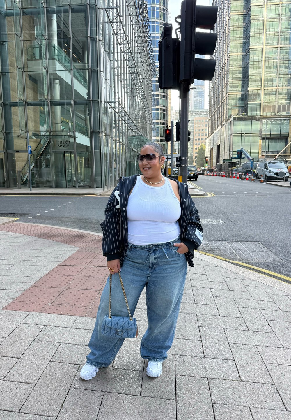 Uma pessoa vestindo uma t-shirt branca, um casaco preto riscado e jeans de perna larga azul claro, segurando uma bolsa cinza acolchoada. Ténis brancos.