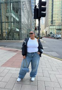 Uma pessoa vestindo uma t-shirt branca, um casaco preto riscado e jeans de perna larga azul claro, segurando uma bolsa cinza acolchoada. Ténis brancos.