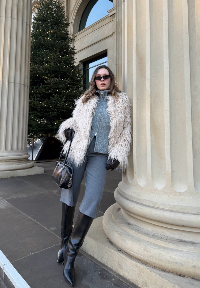 Veste en fausse fourrure crème, pull ajusté gris, pantalon gris coupé court, bottes noires montantes jusqu'aux genoux et petit sac à main noir, debout près d'une colonne.