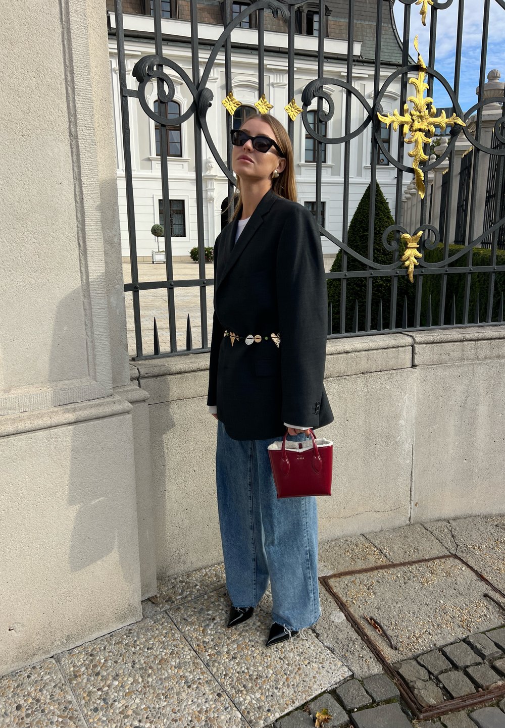 Blazer noir oversize, chemise blanche, jeans taille haute bleu clair, chaussures noires à bout pointu, tenant un petit sac à main rouge avec des accents argentés.